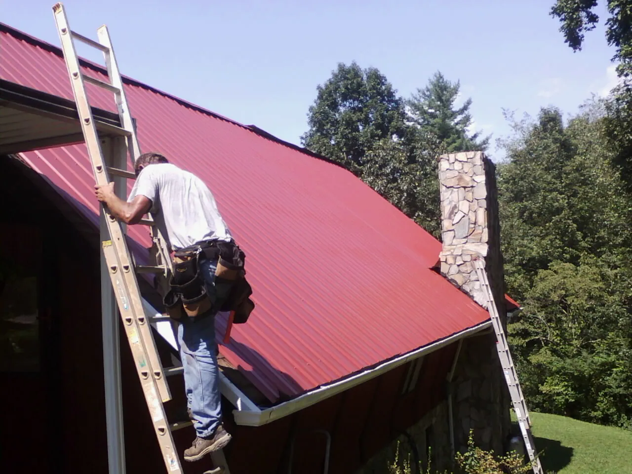 Roofer on a ladder beside a red metal roof and stone chimney — Soffit & Fascia Repair by Roofing Brothers in New Tazewell
